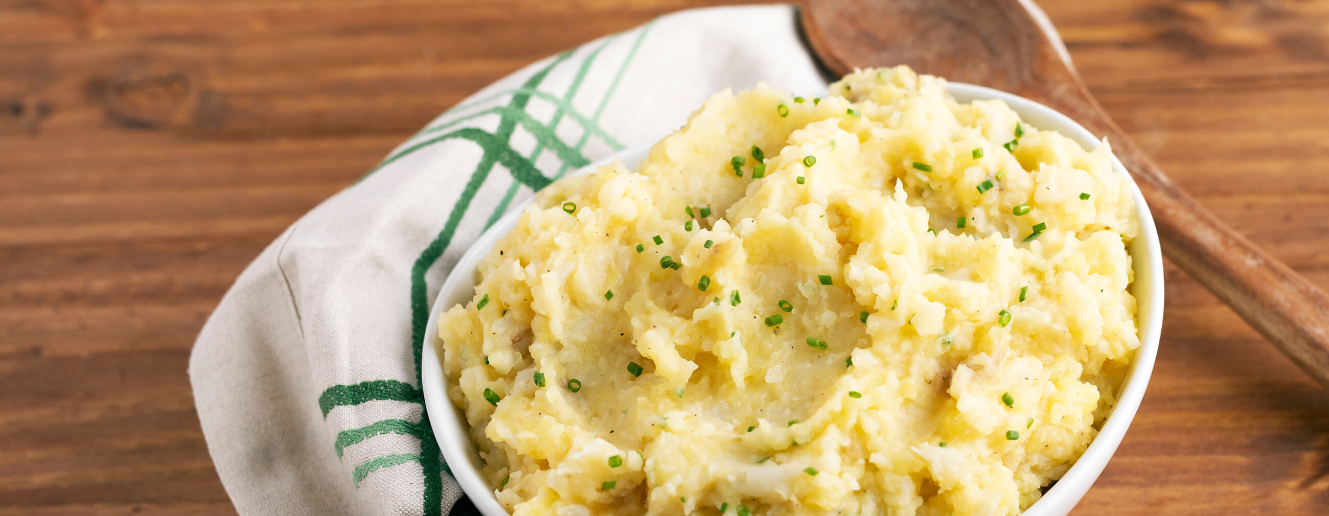 Steaming Cauliflower Mashed Potatoes garnished with chives in a white bowl on a wood table with white cloth napkin and spoon.